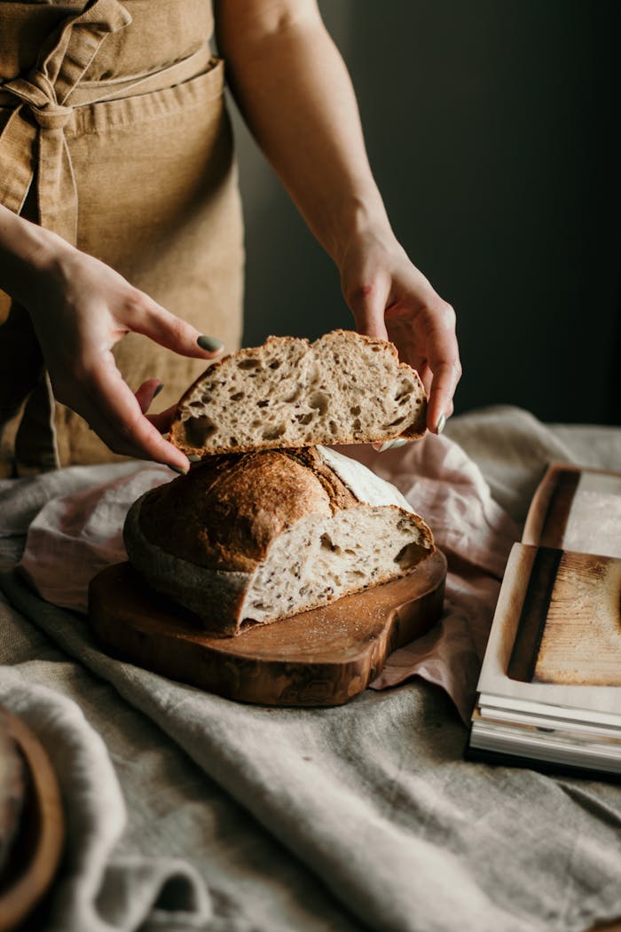 High angle of crop anonymous female baker in apron with piece of bread made with recipe from book at home